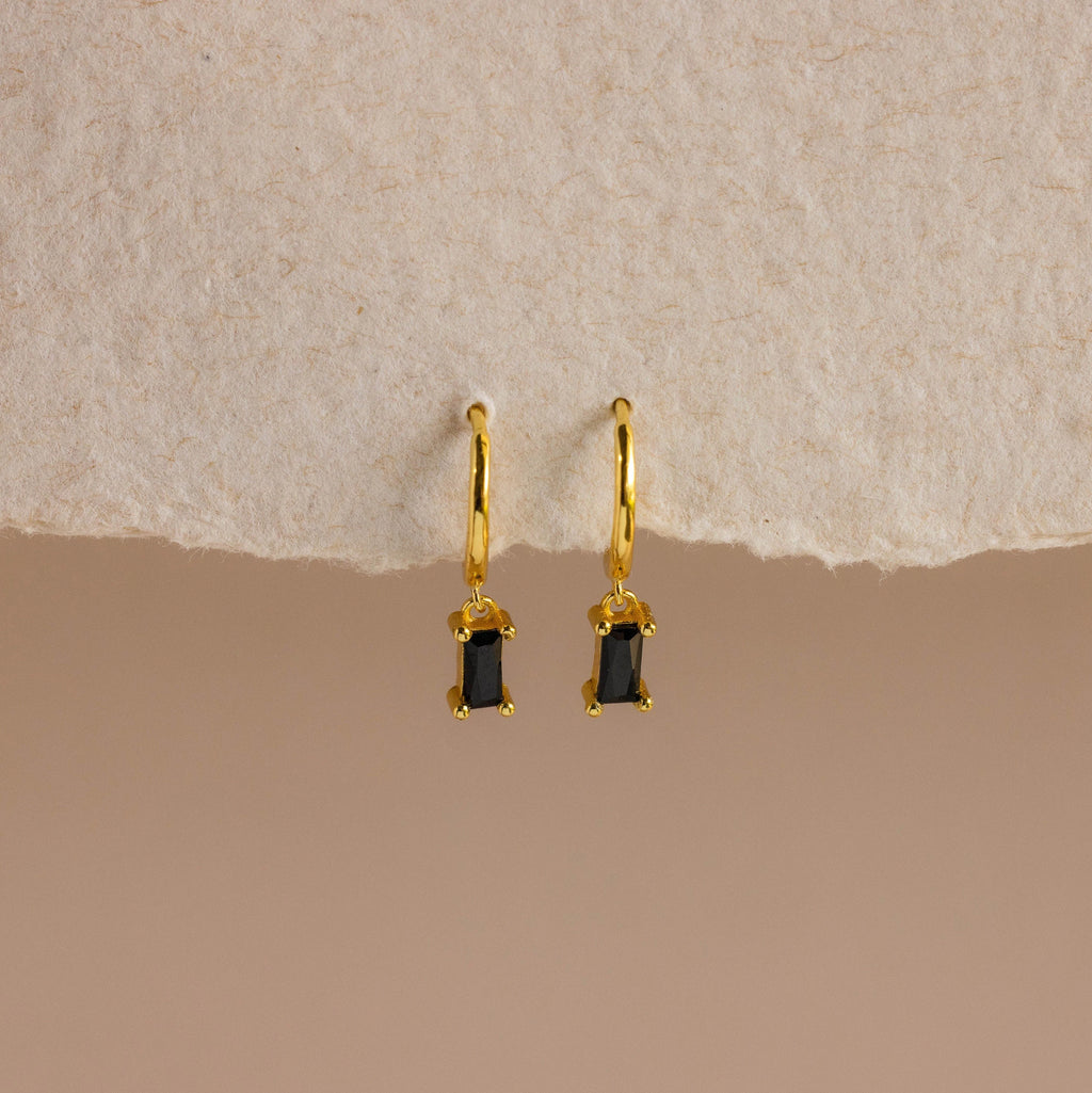 Onyx Baguette Drop Huggies—gold huggie hoops with rectangular onyx baguette drop pendants—shown on a textured beige background.
