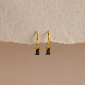 Onyx Baguette Drop Huggies—gold huggie hoops with rectangular onyx baguette drop pendants—shown on a textured beige background.