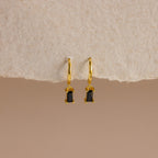 Onyx Baguette Drop Huggies—gold huggie hoops with rectangular onyx baguette drop pendants—shown on a textured beige background.