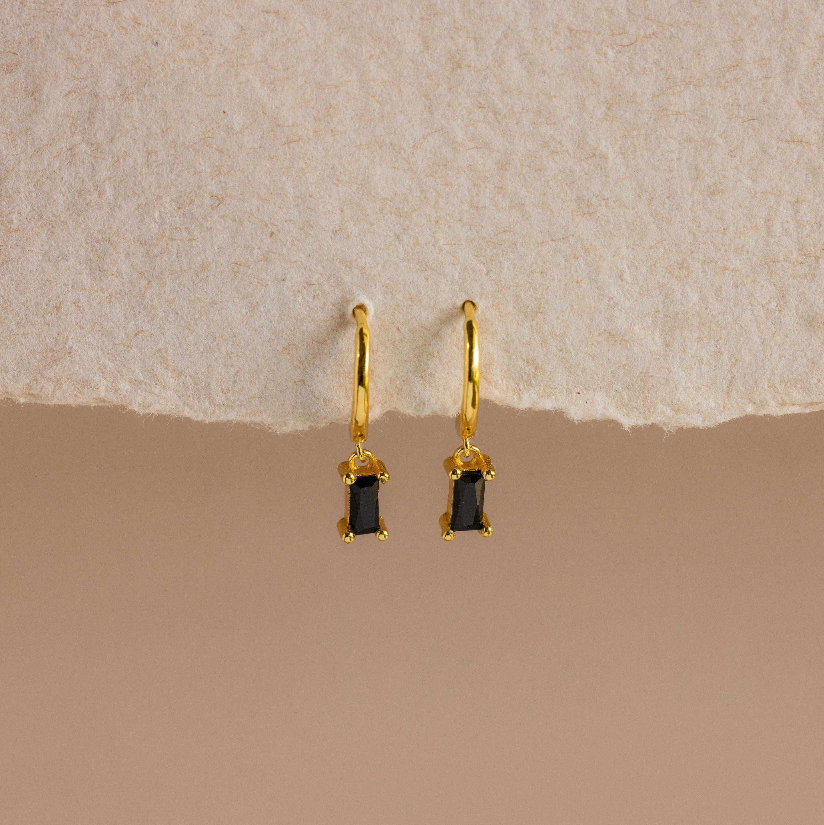 Onyx Baguette Drop Huggies—gold huggie hoops with rectangular onyx baguette drop pendants—shown on a textured beige background.