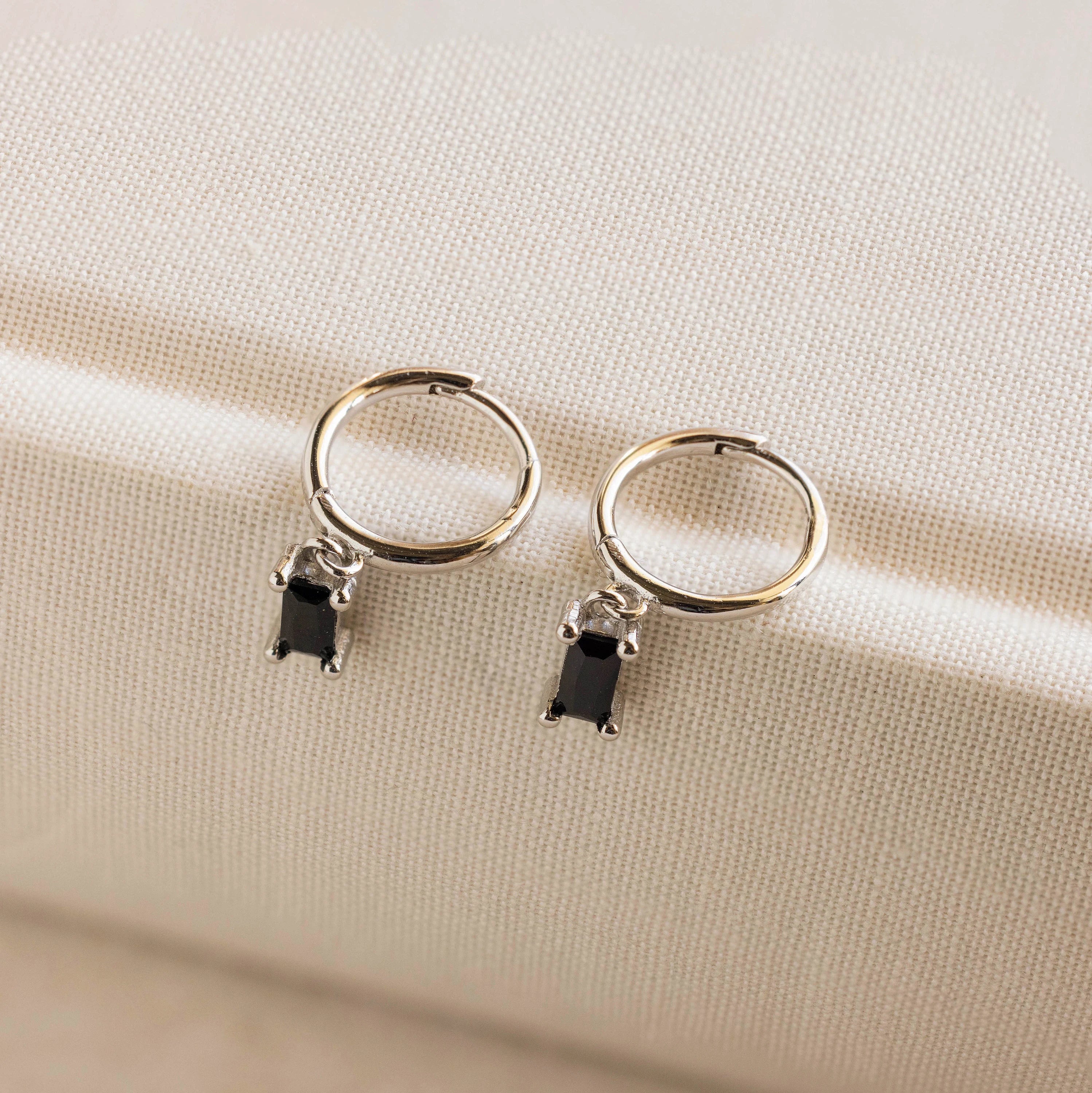Onyx Baguette Drop Huggies featuring silver hoops with small rectangular onyx stone charms, displayed on a cream fabric surface.