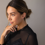 A woman with brown hair in a low bun, dressed in a sheer black top, gazes thoughtfully to the side while wearing Smokey Pearl Drop Huggies earrings.