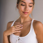 Woman in a white tank top wears gold jewelry, including the Pave Onyx Heart Necklace in Sterling Silver and rings, with her hand on her chest.