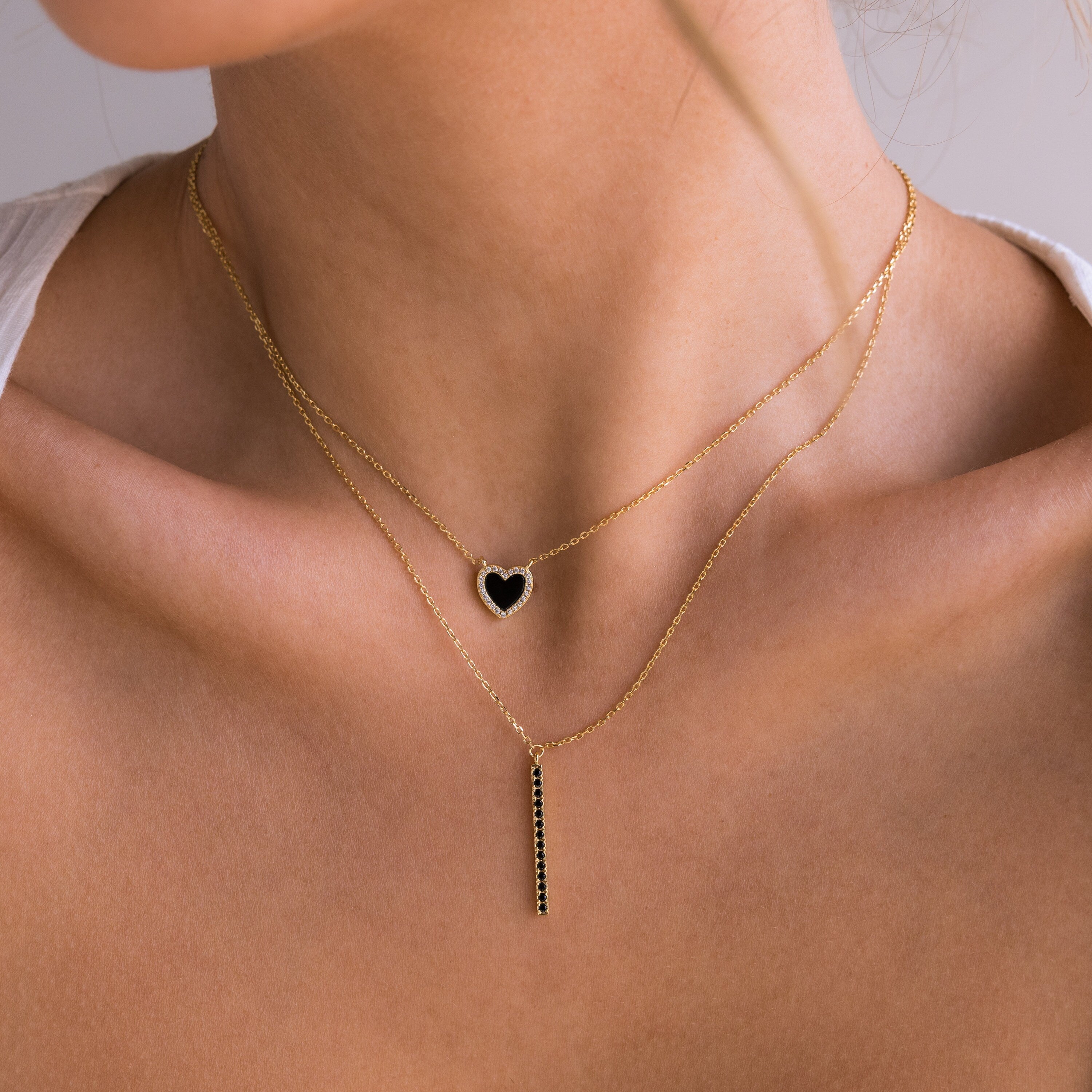 A woman wears two gold necklaces: the Pave Onyx Heart Necklace and a charm necklace with a vertical black bar pendant.
