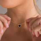 A person holding a Pave Onyx Heart Necklace in 18K Gold, its heart-shaped charm shimmering between their fingers.