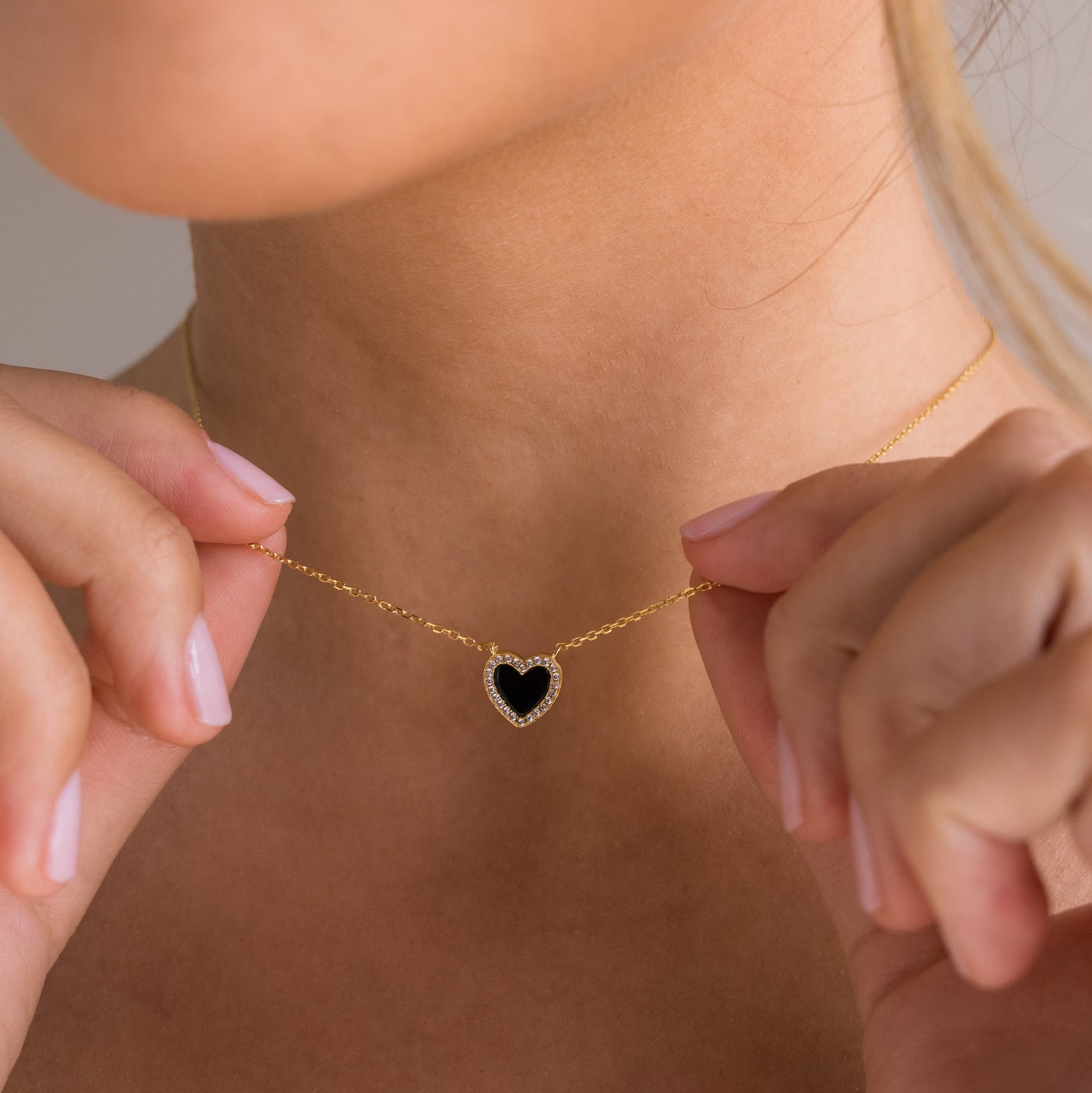 A person holding a Pave Onyx Heart Necklace in 18K Gold, its heart-shaped charm shimmering between their fingers.