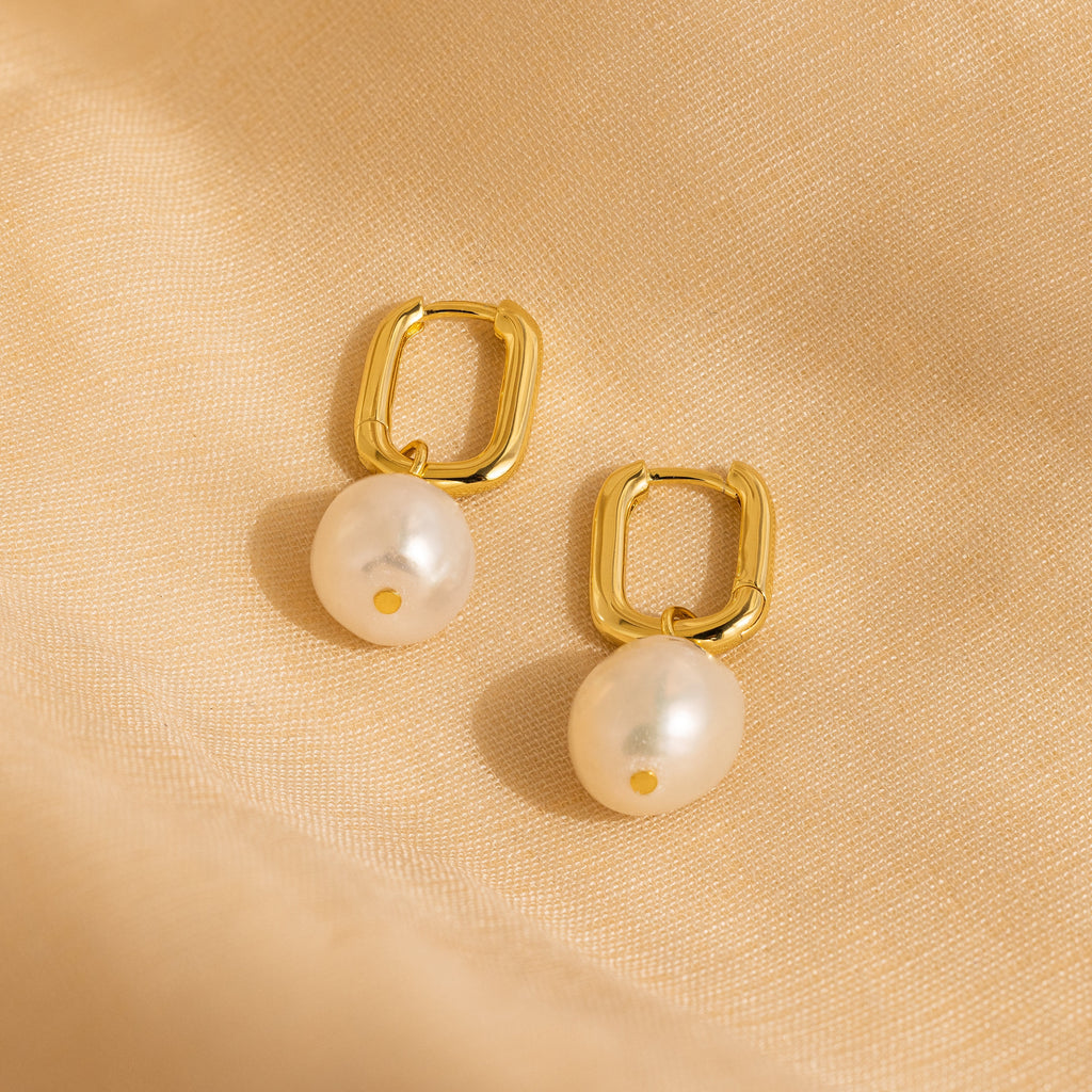 Freshwater Pearl Drop Hoops feature gold hoops with dangling pearls, exuding timeless elegance. Shown on a soft beige fabric, they add a refined touch to any look.