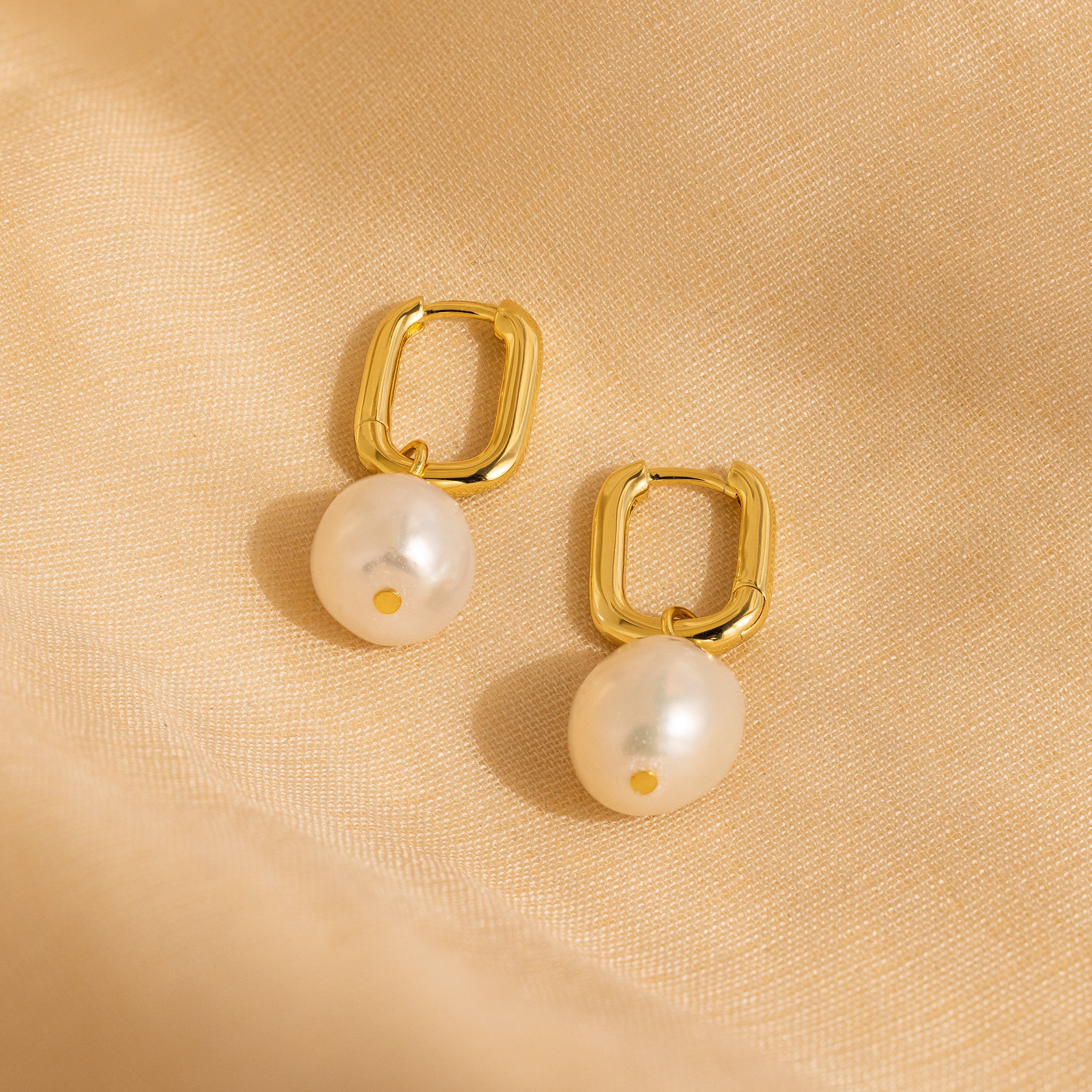 Freshwater Pearl Drop Hoops feature gold hoops with dangling pearls, exuding timeless elegance. Shown on a soft beige fabric, they add a refined touch to any look.