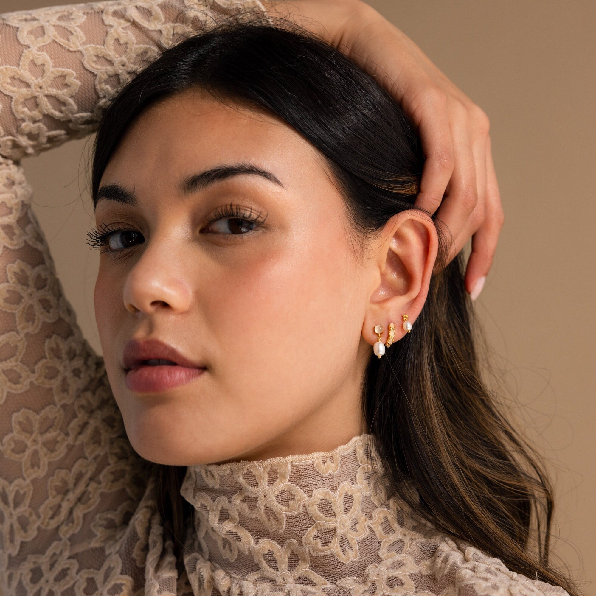 A woman with long dark hair in a lace top wears Moonstone Pearl Drop Earrings, touching her ear and gazing at the camera.