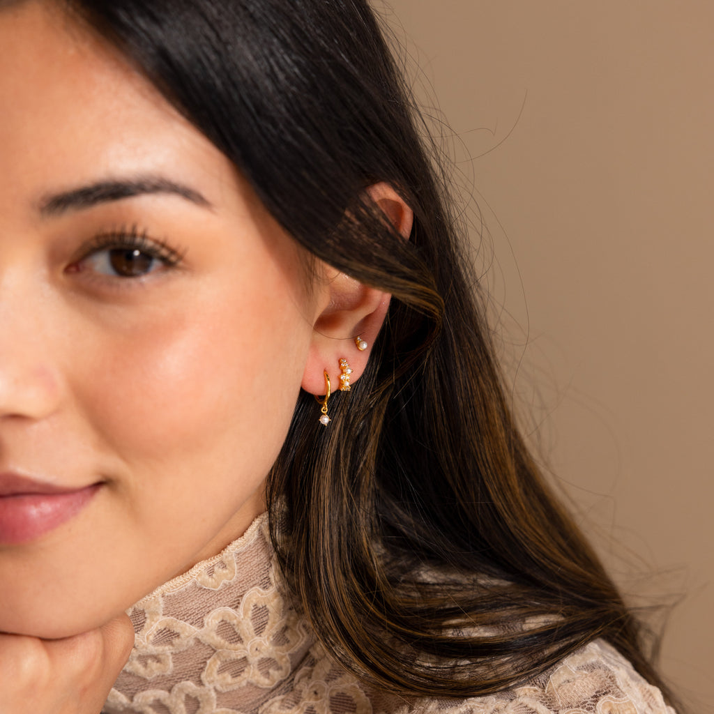 A woman wearing Pearl Diamond Cluster Huggies in 18K Gold and a lace high-neck top rests her chin on her hand against a neutral background.