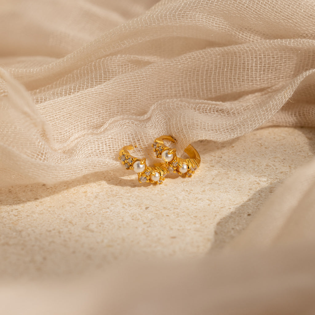 The Pearl Diamond Cluster Huggies in 18K Gold, featuring a floral-inspired pearl and diamond design, rest on a beige textured surface with sheer fabric—ideal for those seeking elegant pearl earrings.