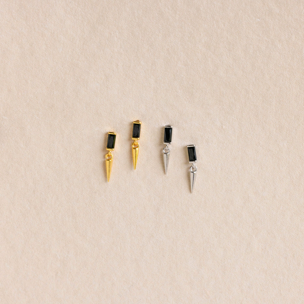 Two pairs of Onyx Dagger Drop Earrings—one gold, one silver—each with baguette-cut onyx stones in a sleek rectangular design, displayed on a beige background.