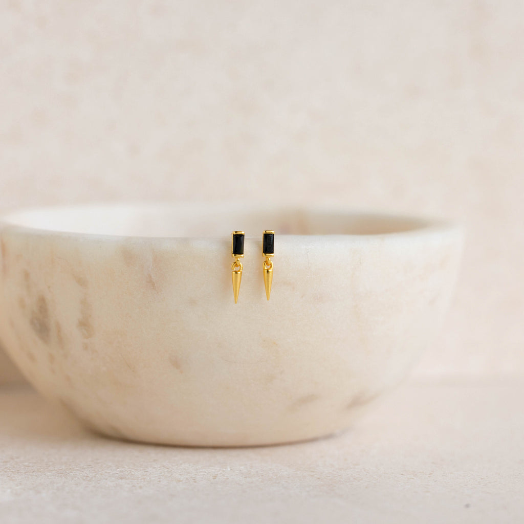 Onyx Dagger Drop Earrings in gold and black are displayed on the rim of a white marble bowl.