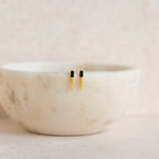 Onyx Dagger Drop Earrings in gold and black are displayed on the rim of a white marble bowl.