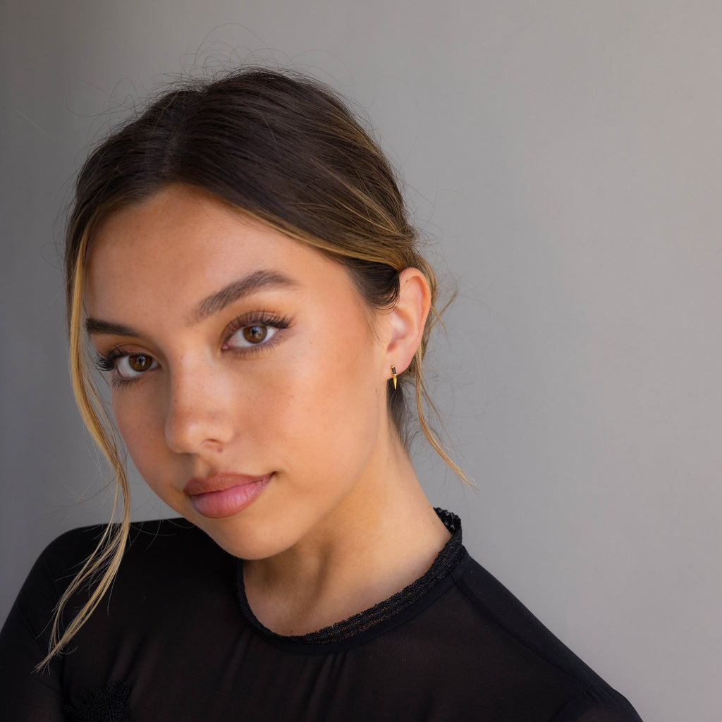 A young woman with brown hair and subtle makeup wears a black top and the Onyx Dagger Drop Earrings, each featuring a delicate dagger charm, as she poses against a plain gray background.