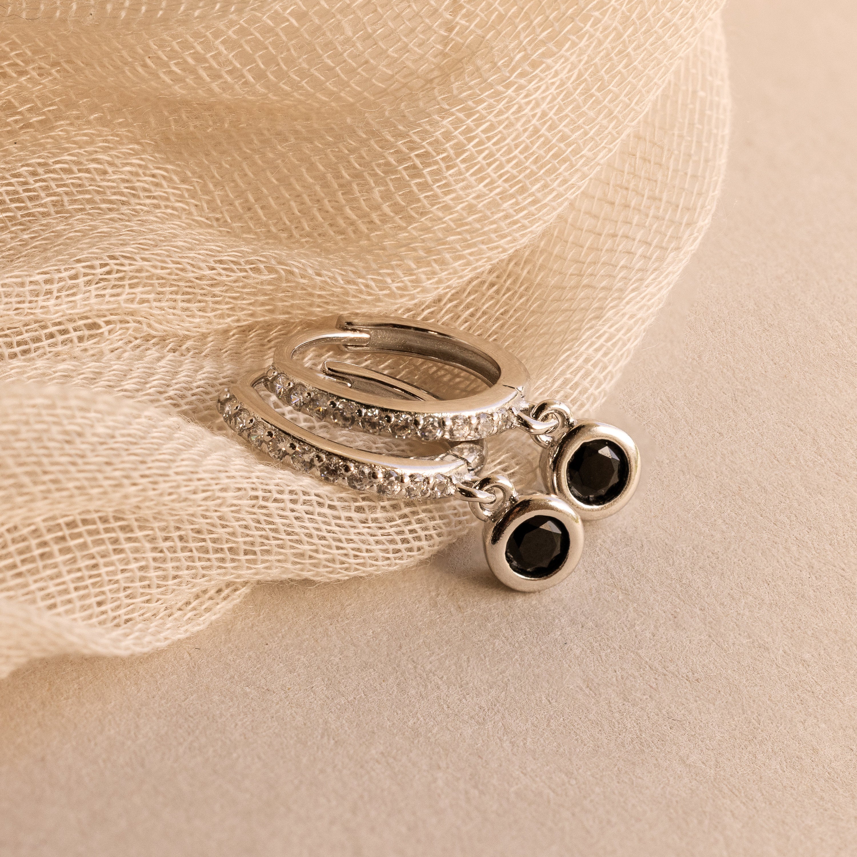 Pave Onyx Drop Huggies: silver hoops adorned with black onyx gemstone drops and delicate diamonds, displayed on soft beige fabric.