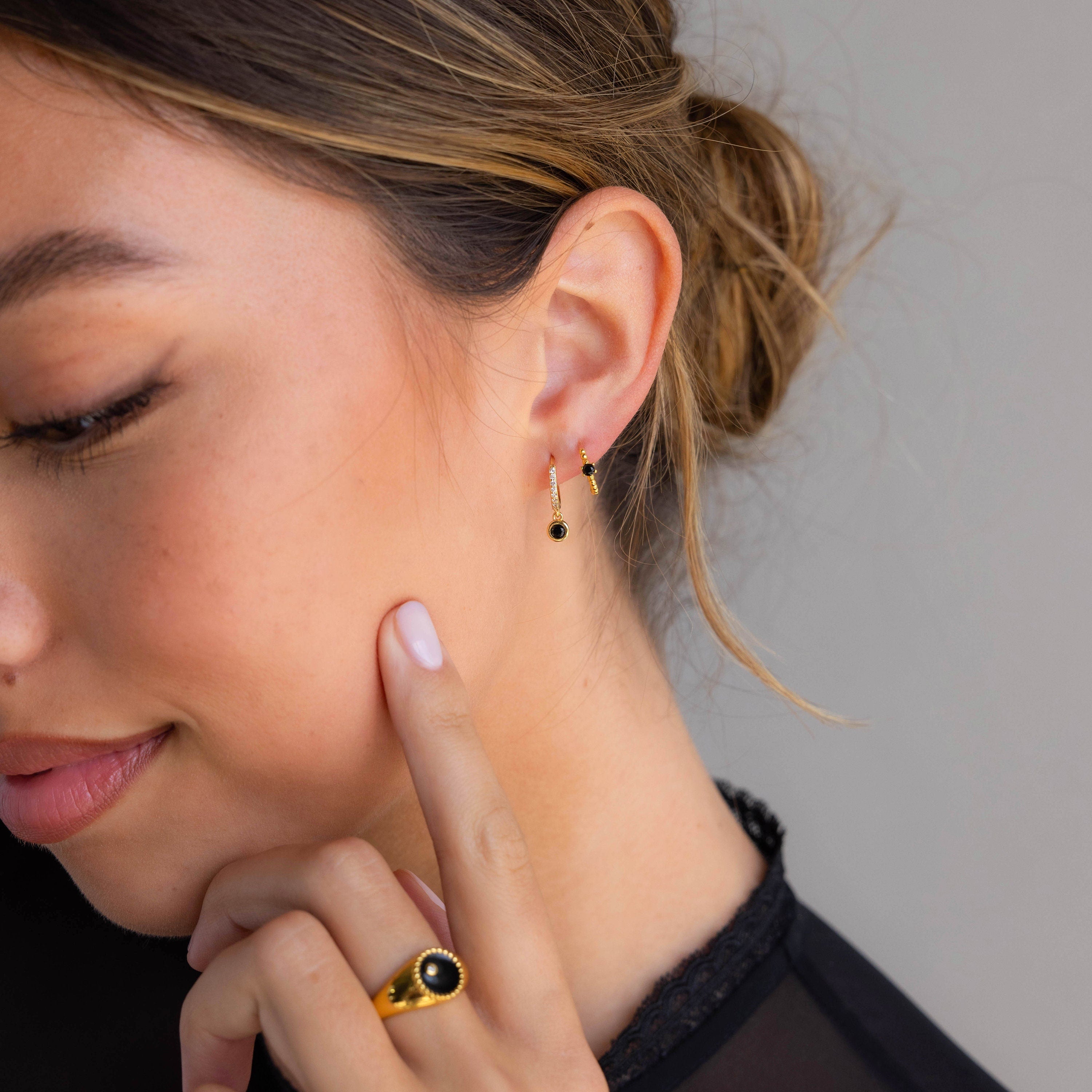A woman with a low bun wears gold earrings and a matching hexagonal ring, touching her cheek, featuring the Onyx Beaded Huggies.