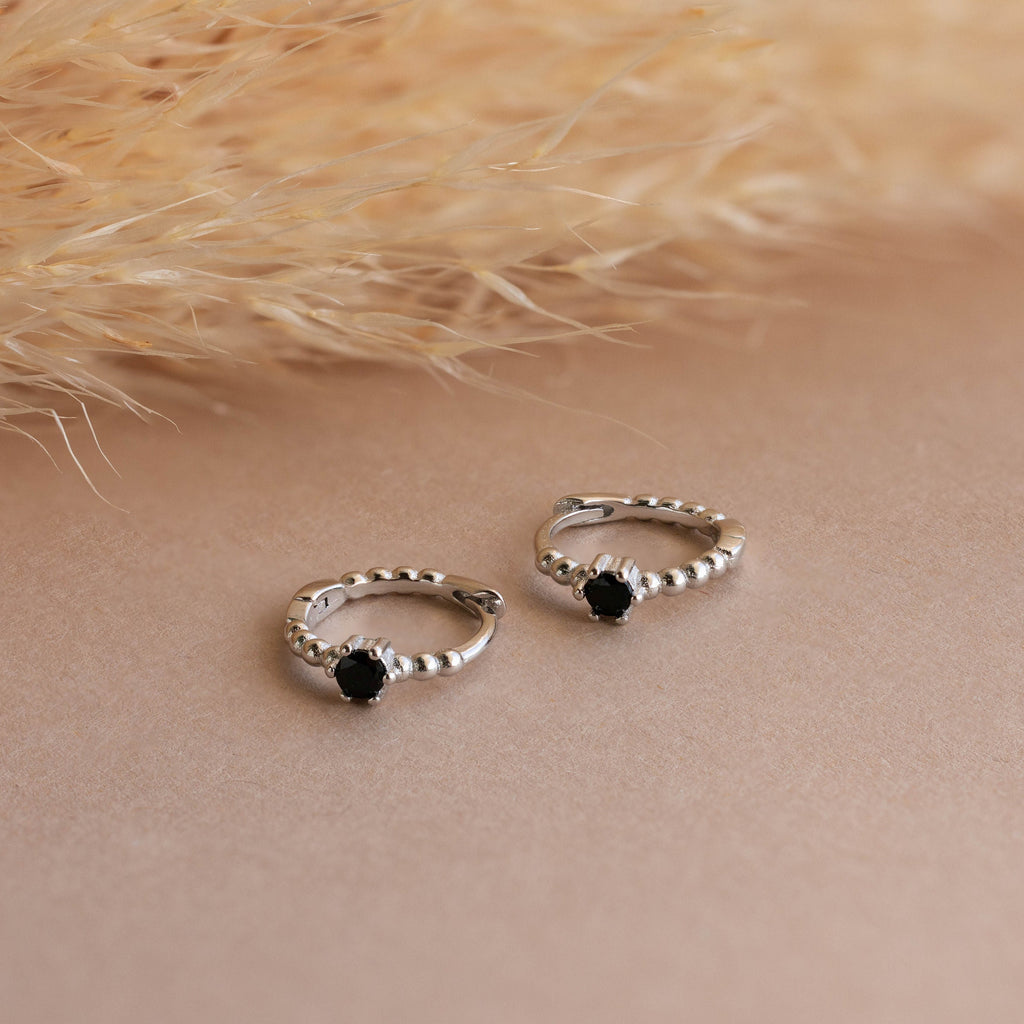 Onyx Beaded Huggies with black stones are displayed on a neutral surface beside dried grass, lending an elegant touch to any look.