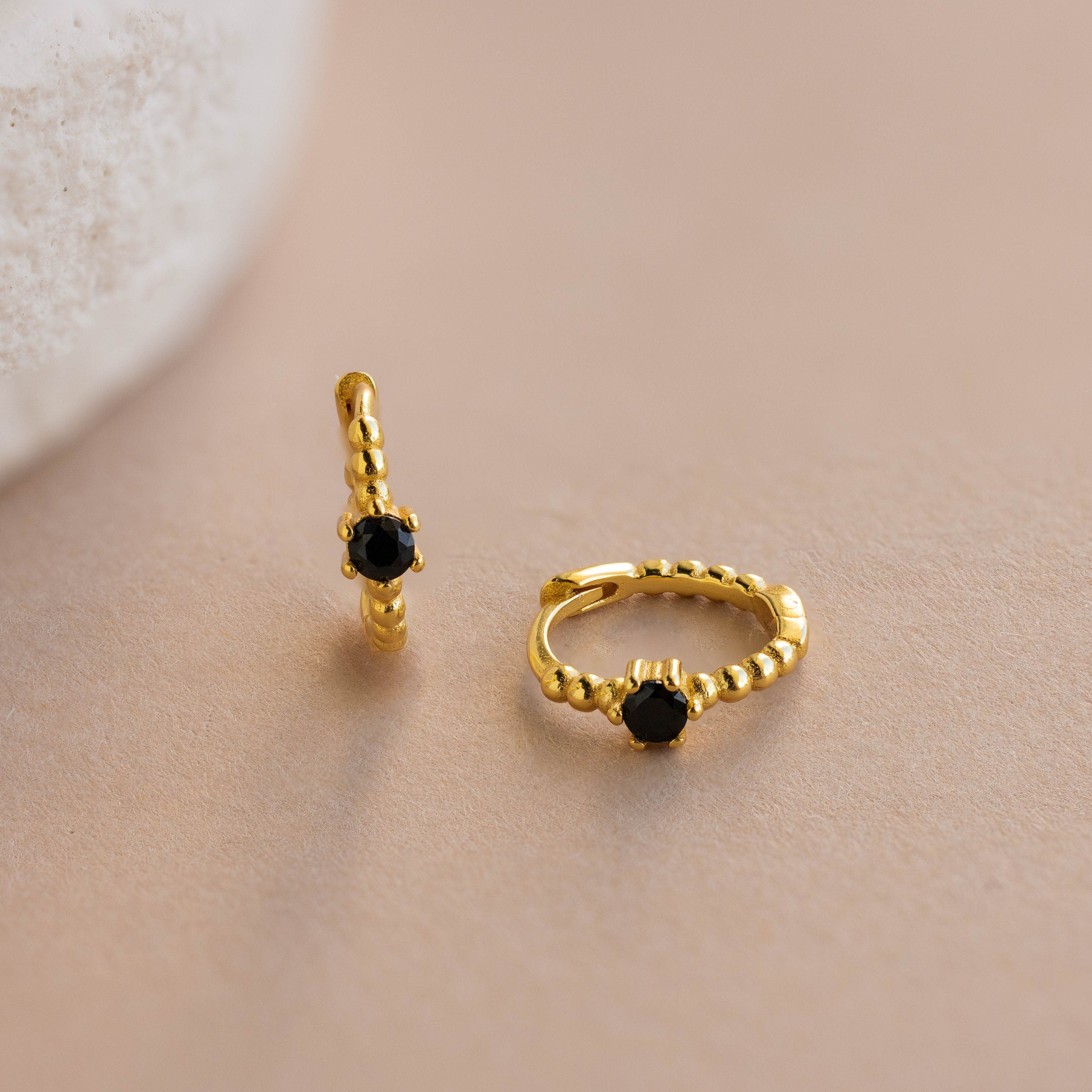 Two small Onyx Beaded Huggies featuring a beaded gold texture and black stones, displayed on a beige surface.