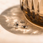Tiny Onyx Pearl Bar Studs, featuring black and clear stones, are displayed next to a ribbed glass on a sunlit surface for a chic look.