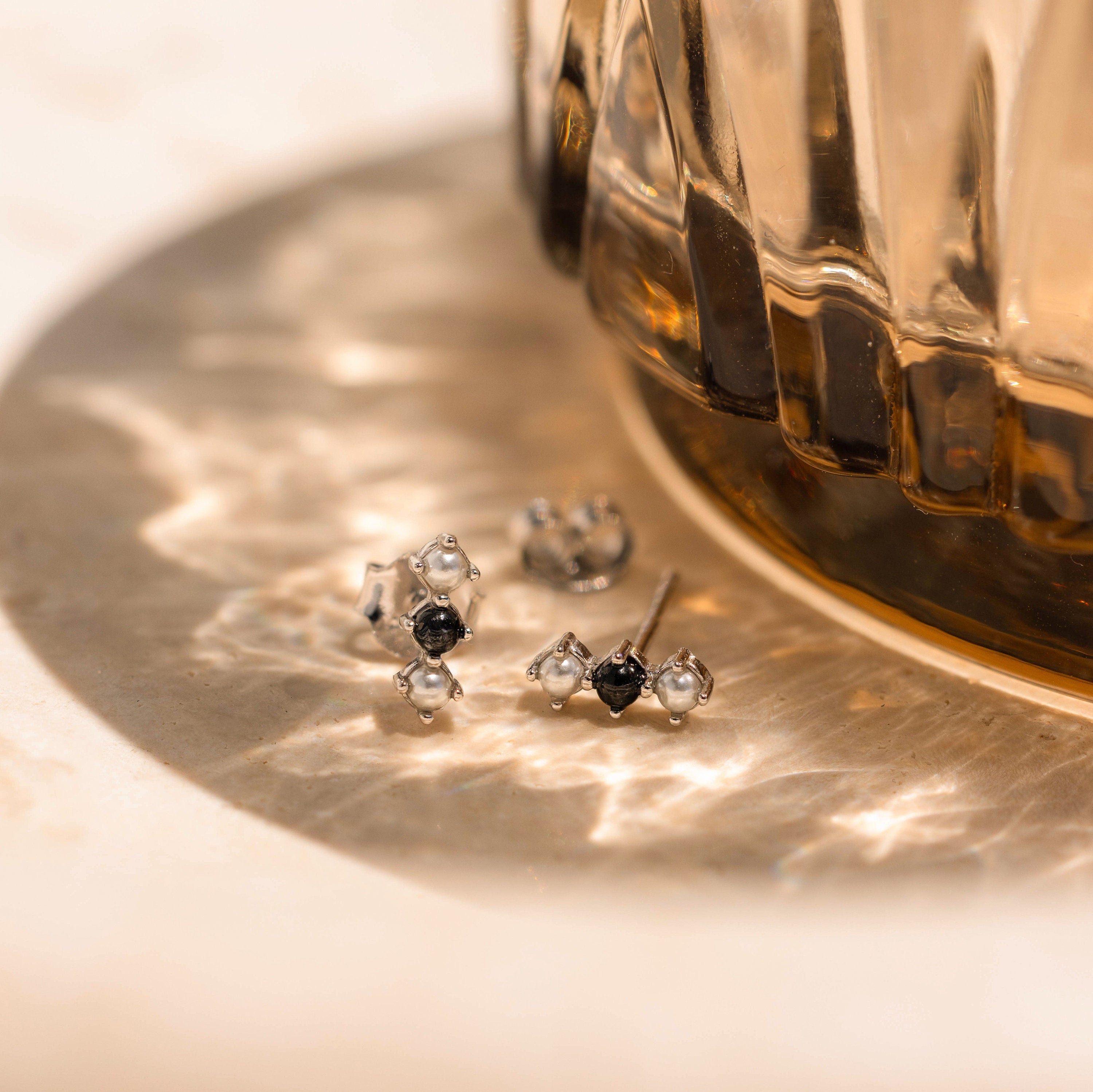 Tiny Onyx Pearl Bar Studs, featuring black and clear stones, are displayed next to a ribbed glass on a sunlit surface for a chic look.