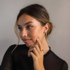 Woman with dark hair in a low bun, wearing a black top and elegant Tiny Onyx Pearl Bar Studs, touches her chin and smiles softly.
