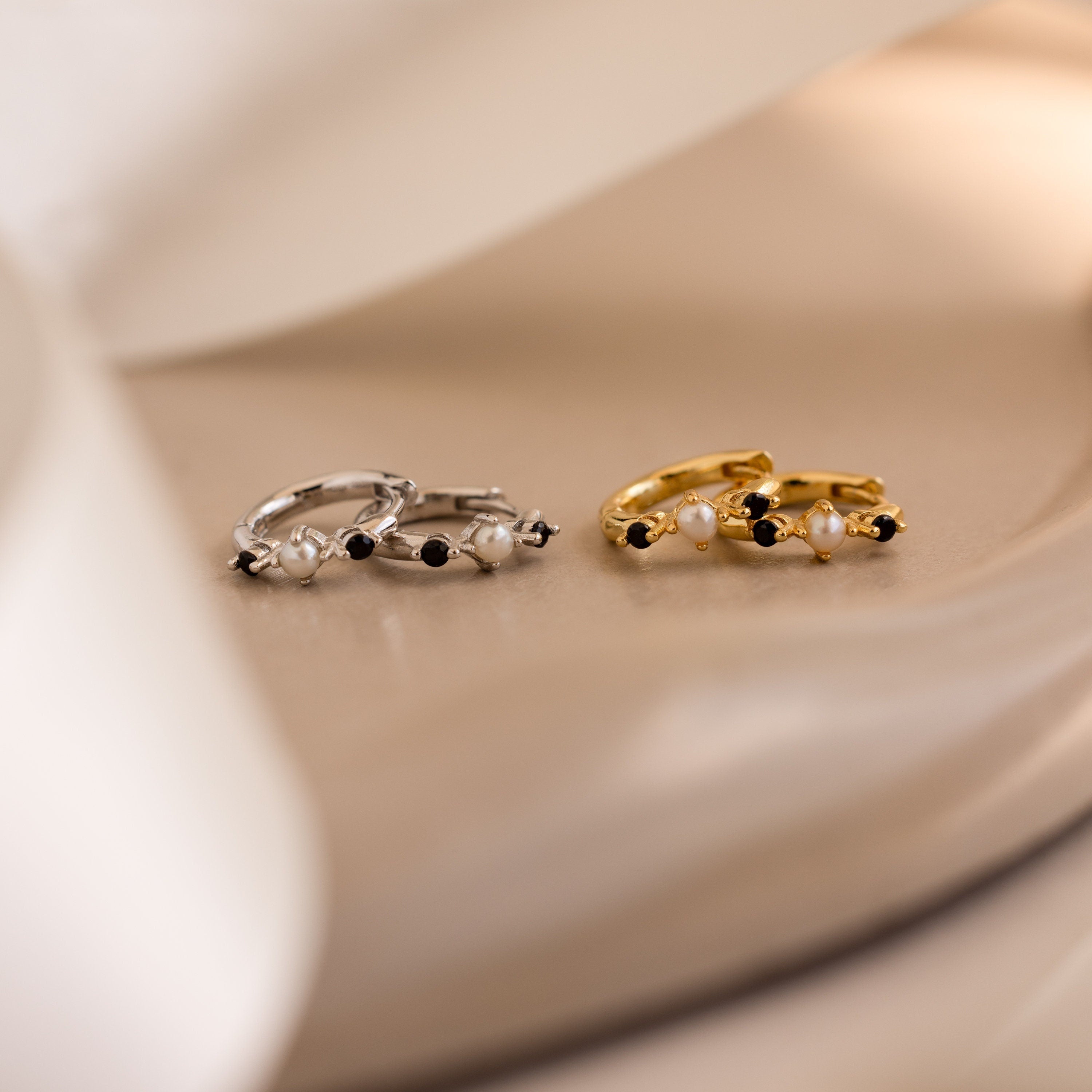 The Pearl & Onyx Huggies—one pair in silver, one in gold—feature pearls and black stones for a chic, elegant look on a beige surface, ideal for anyone seeking timeless pearl earrings.