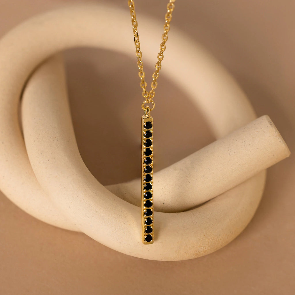 The Onyx Bar Pendant Necklace features a gold chain and vertical bar with onyx gemstones, shown on a beige abstract background—a modern piece ideal for daily elegance.