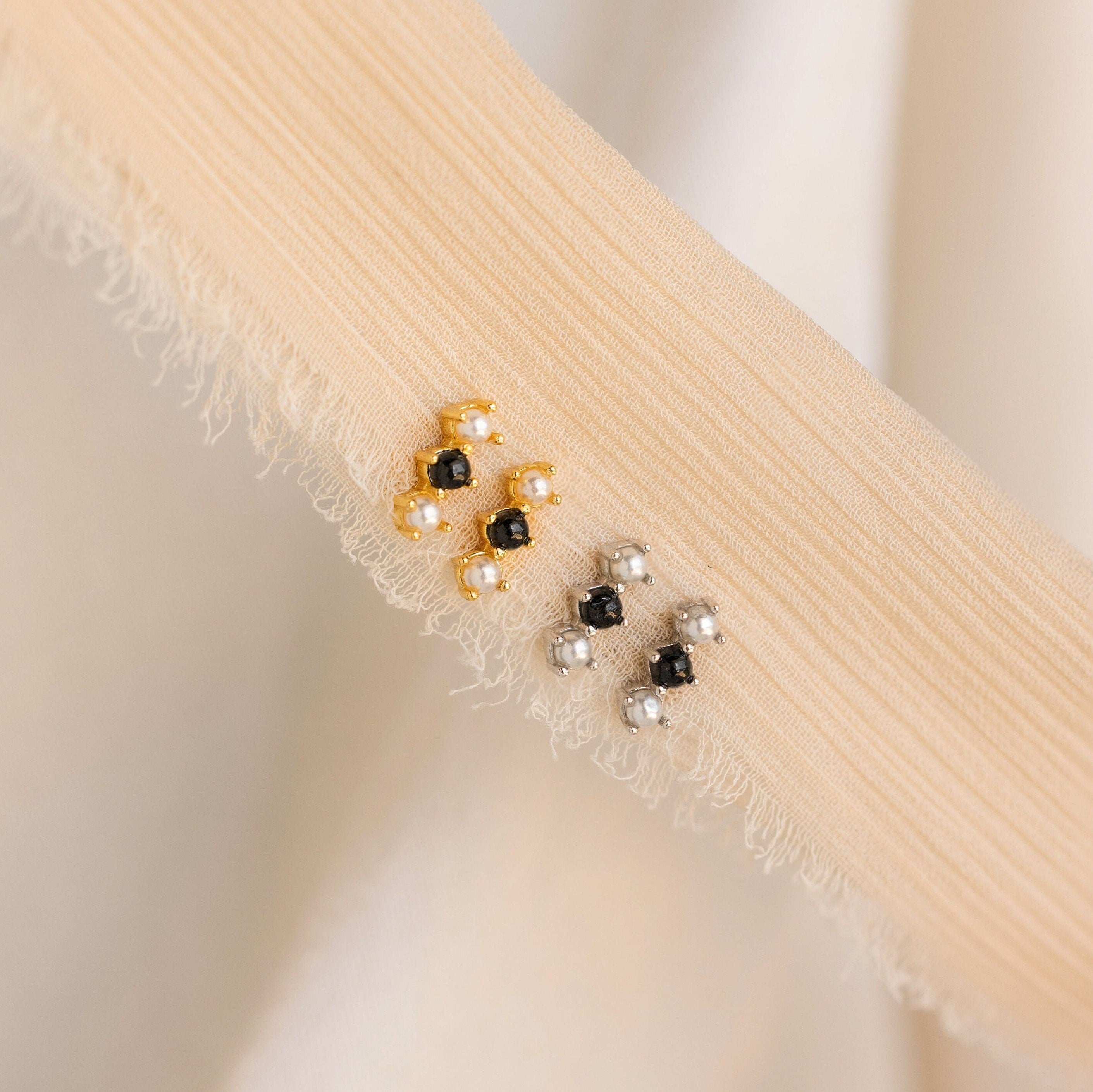 Two pairs of Tiny Onyx Pearl Bar Studs—one gold, one silver—are displayed on textured beige fabric, bringing a modern touch to your everyday style.