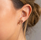 A close-up of a woman's ear with two small gold studs, hair tied back and a neutral background. Add our Pearl & Onyx Huggies for timeless elegance.