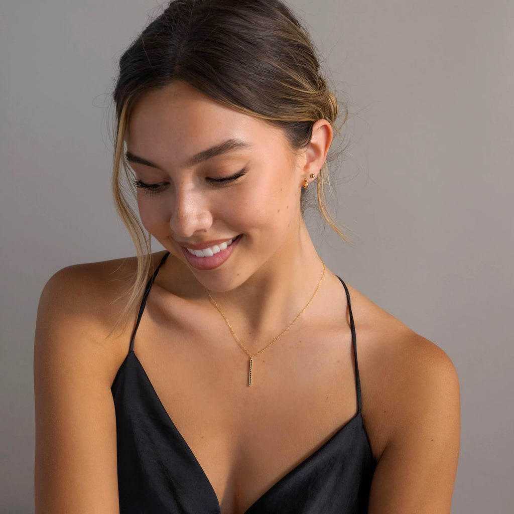 A woman in a black dress smiles and looks down, her elegant style accentuated by small earrings and the Onyx Bar Pendant Necklace, which features a sleek bar design highlighted with refined onyx gemstones.