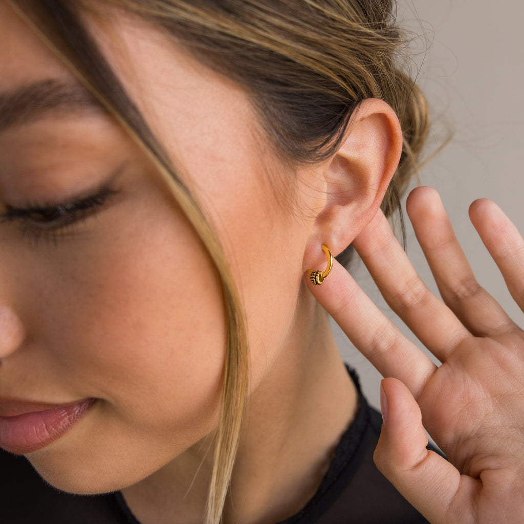 A woman with light brown hair displays the Onyx Pave Beaded Hoops, holding her hand near her ear to highlight the delicate gold earrings.
