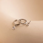 The Diamond Pave Earrings Set, featuring two silver hoop earrings with square diamond pendants and one stud earring, rests on a smooth beige surface—an elegant choice for bridal jewelry.