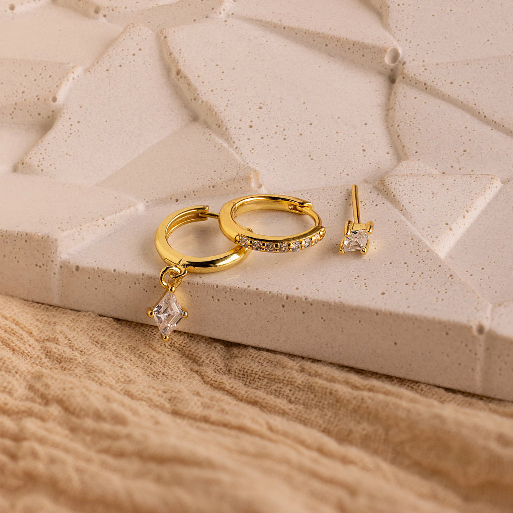 The Diamond Pave Earrings Set features gold hoops with small gemstones and a matching stud, displayed on a textured beige surface—ideal as wedding earrings or elegant bridal jewelry.