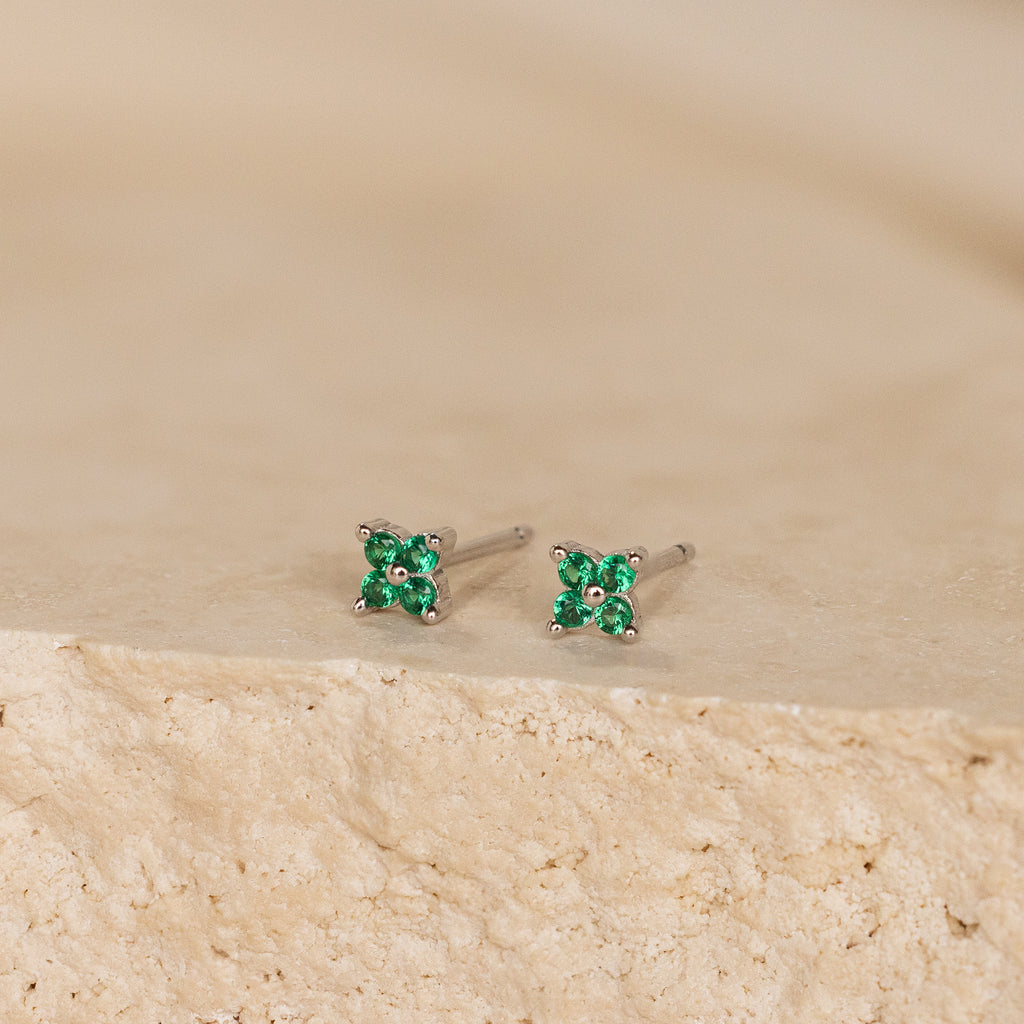 The Emerald Ivy Flower Studs are silver earrings with small green gemstones, reflecting nature-inspired elegance. Shown on a textured beige surface.