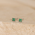 The Emerald Ivy Flower Studs are silver earrings with small green gemstones, reflecting nature-inspired elegance. Shown on a textured beige surface.