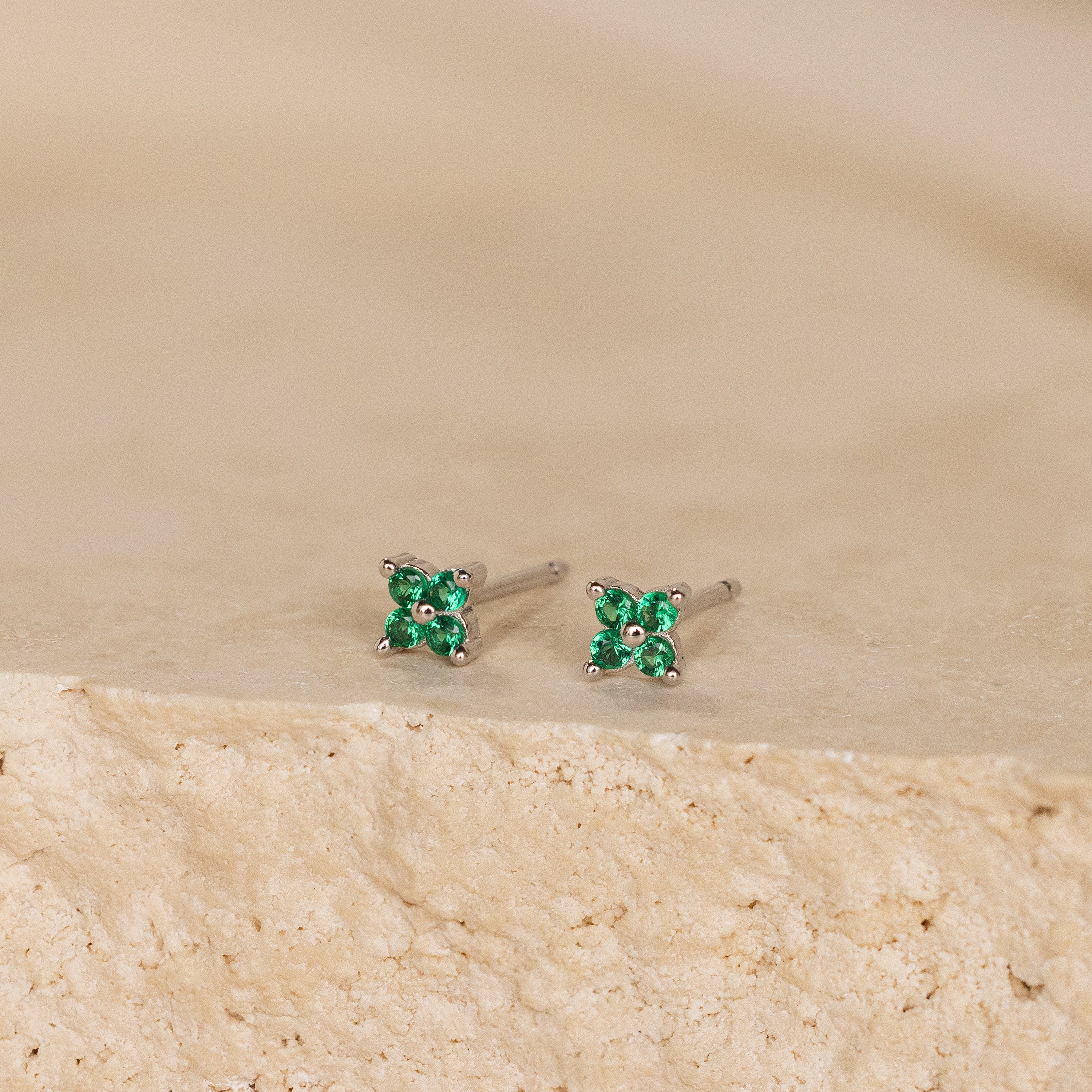 The Emerald Ivy Flower Studs are silver earrings with small green gemstones, reflecting nature-inspired elegance. Shown on a textured beige surface.