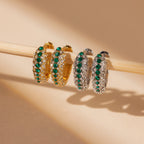 Emerald Pave Snake Hoops—gold hoop earrings with green gemstones—are displayed on a light wooden rod.