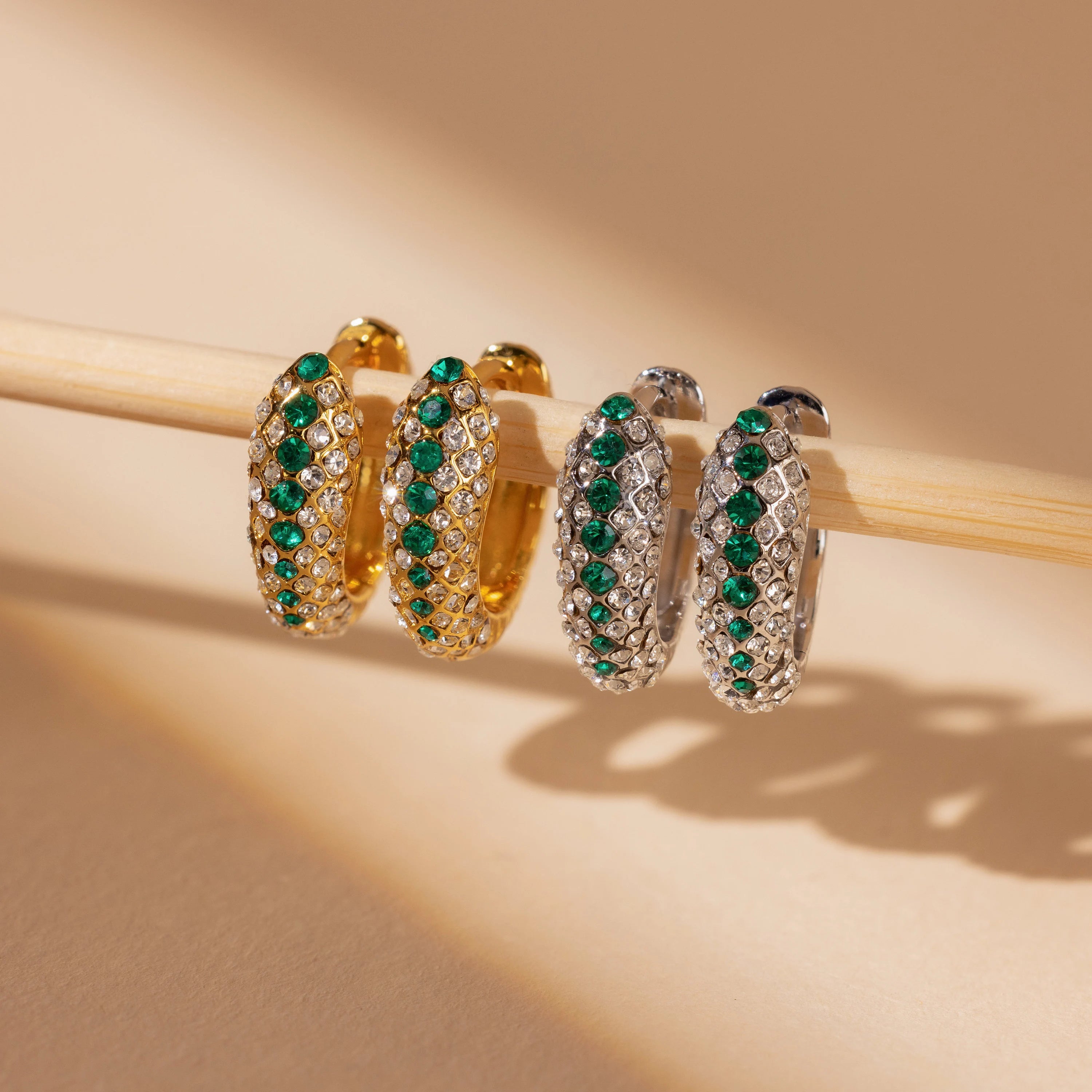Emerald Pave Snake Hoops—gold hoop earrings with green gemstones—are displayed on a light wooden rod.