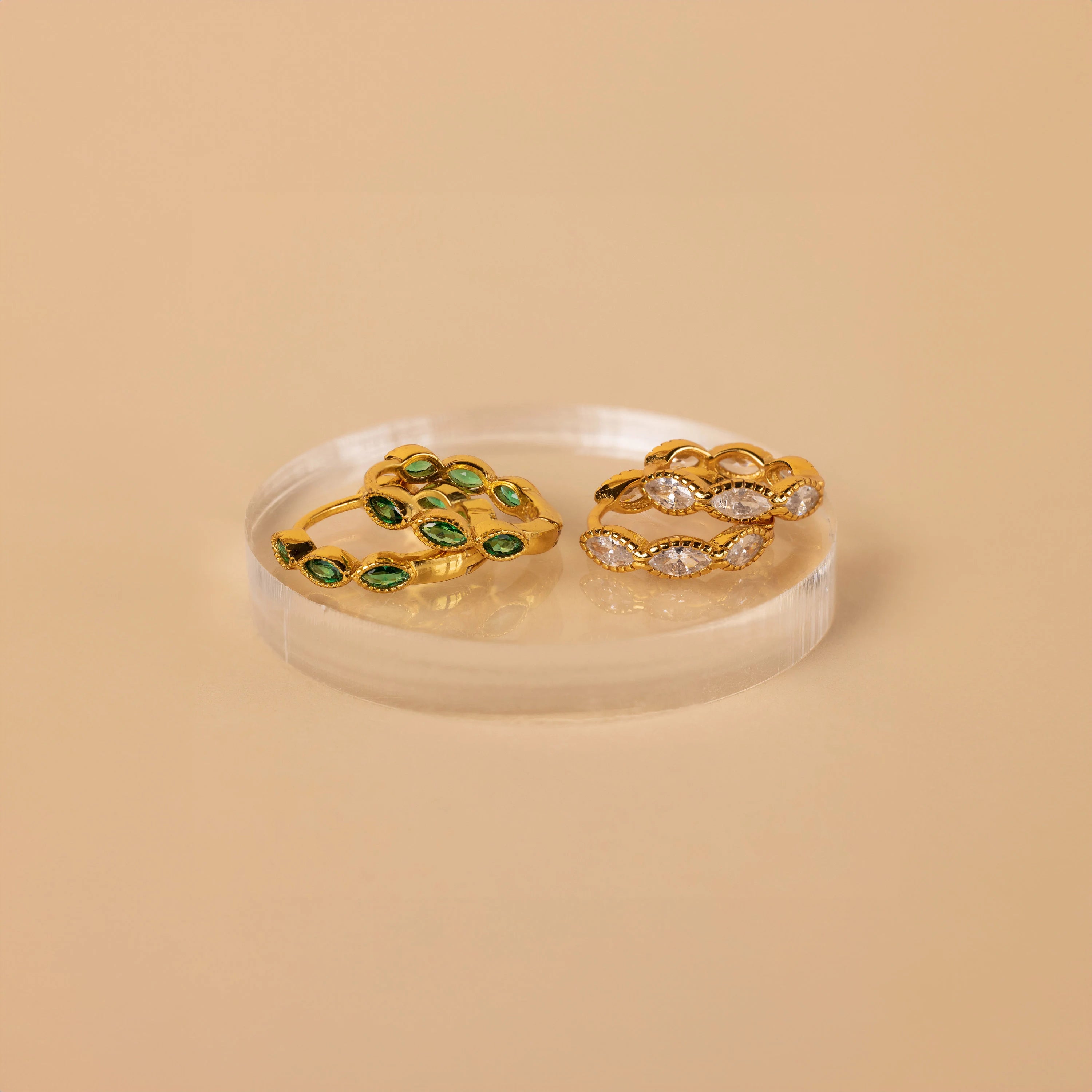 The Emerald Marquise Inlay Huggies, featuring delicate green stones set in gold, are displayed on a round clear stand—perfect dainty earrings to complement your look.