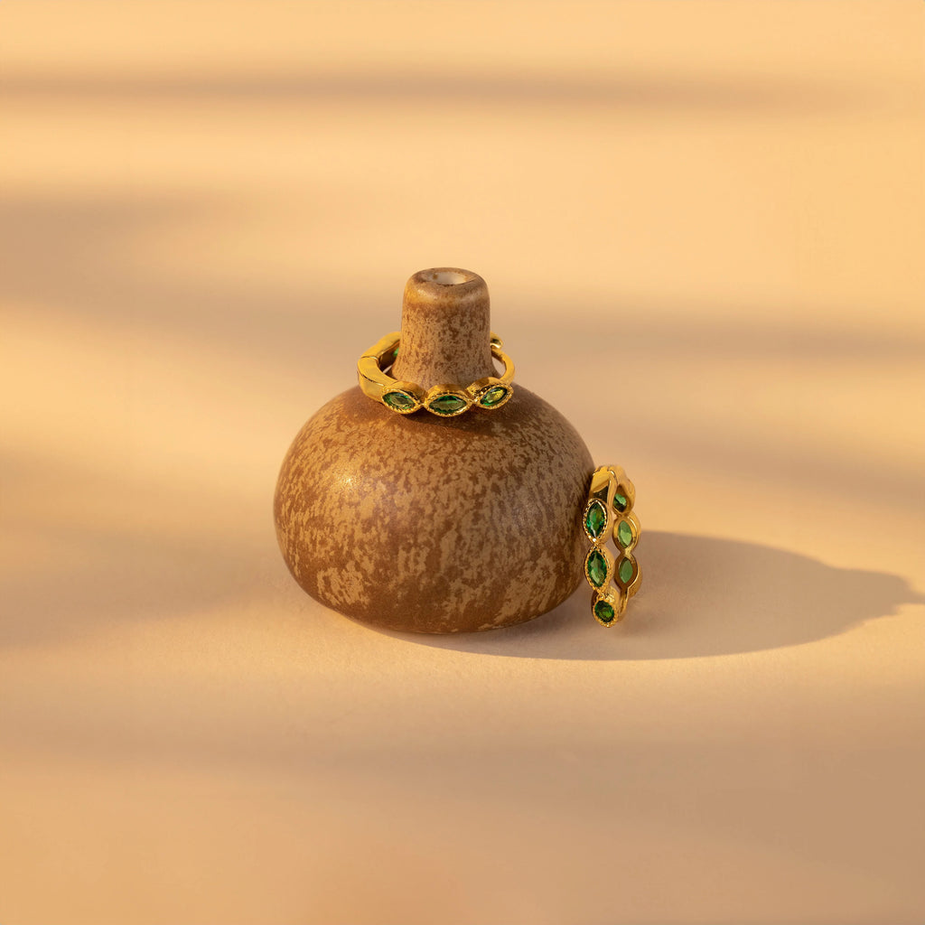Emerald Marquise Inlay Huggies, elegant birthstone earrings with green gemstones, are displayed alongside gold rings and dainty jewelry on a small brown ceramic vase.