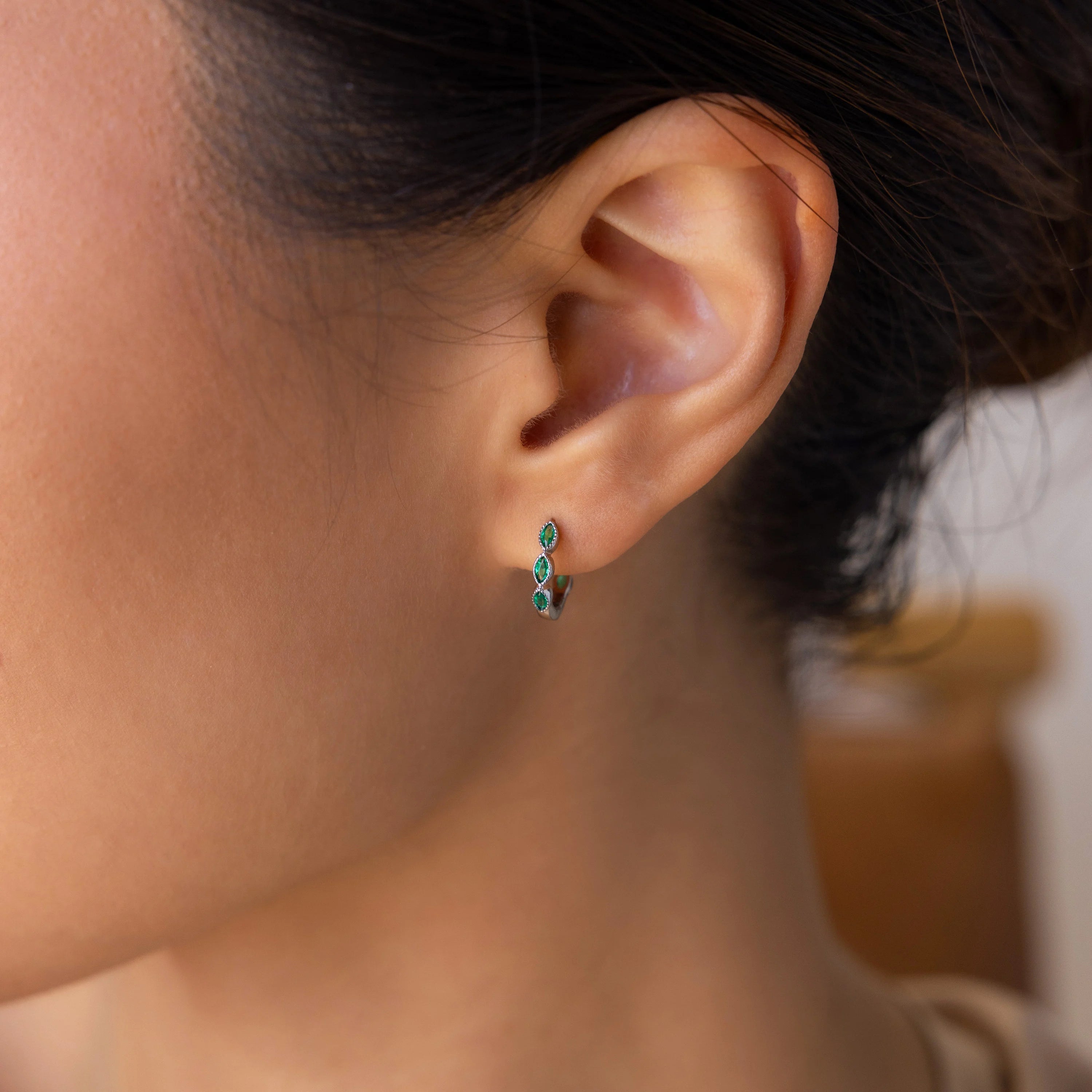 Close-up of a woman’s ear wearing Emerald Marquise Inlay Huggies—dainty silver hoop earrings accented with green gemstones, perfect as birthstone earrings or chic emerald huggies.