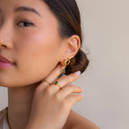 A woman poses with her hand near her face, showcasing Emerald Marquise Inlay Huggies and delicate gold rings against a neutral background.