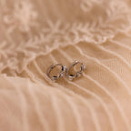 The Ericka Diamond Tennis Huggies sparkle atop soft, cream-colored, embroidered fabric with delicate floral patterns—a scene echoing the timeless elegance of art deco jewelry.