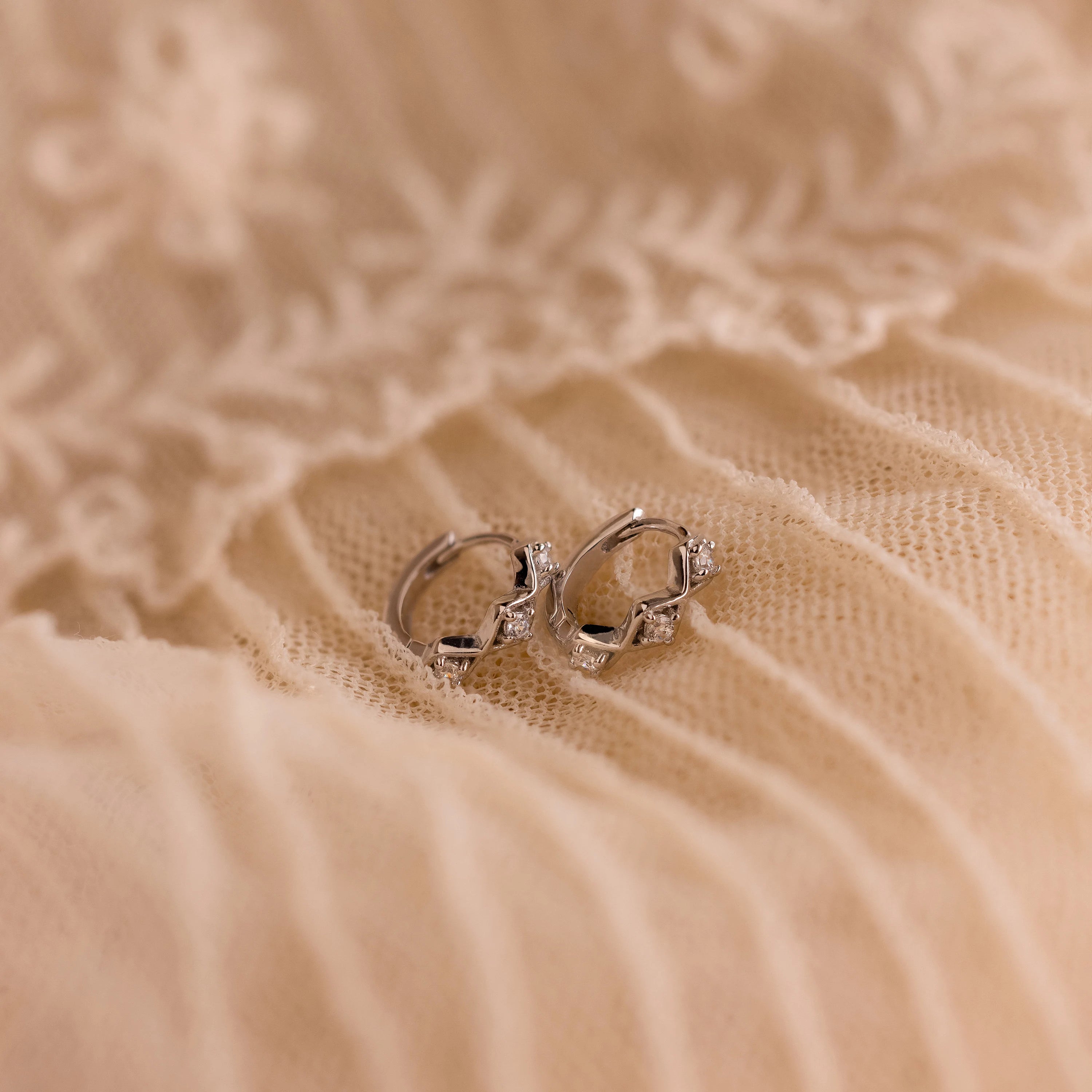 The Ericka Diamond Tennis Huggies sparkle atop soft, cream-colored, embroidered fabric with delicate floral patterns—a scene echoing the timeless elegance of art deco jewelry.