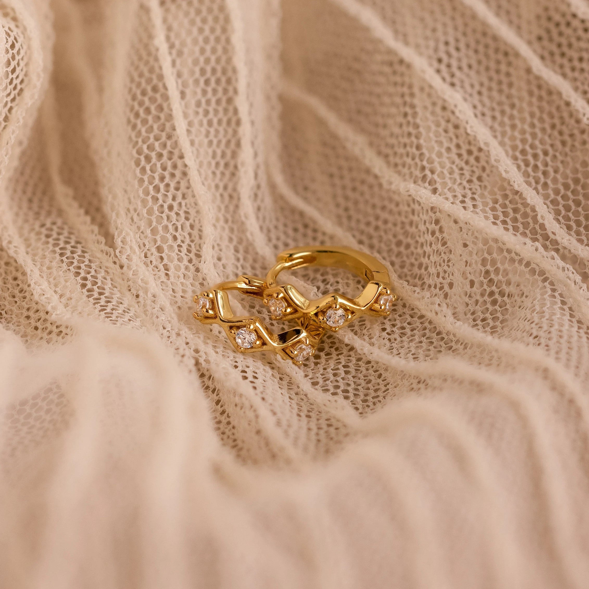 The Ericka Diamond Tennis Huggies are styled with a gold ring on white fabric, creating a timeless and elegant art deco-inspired look.