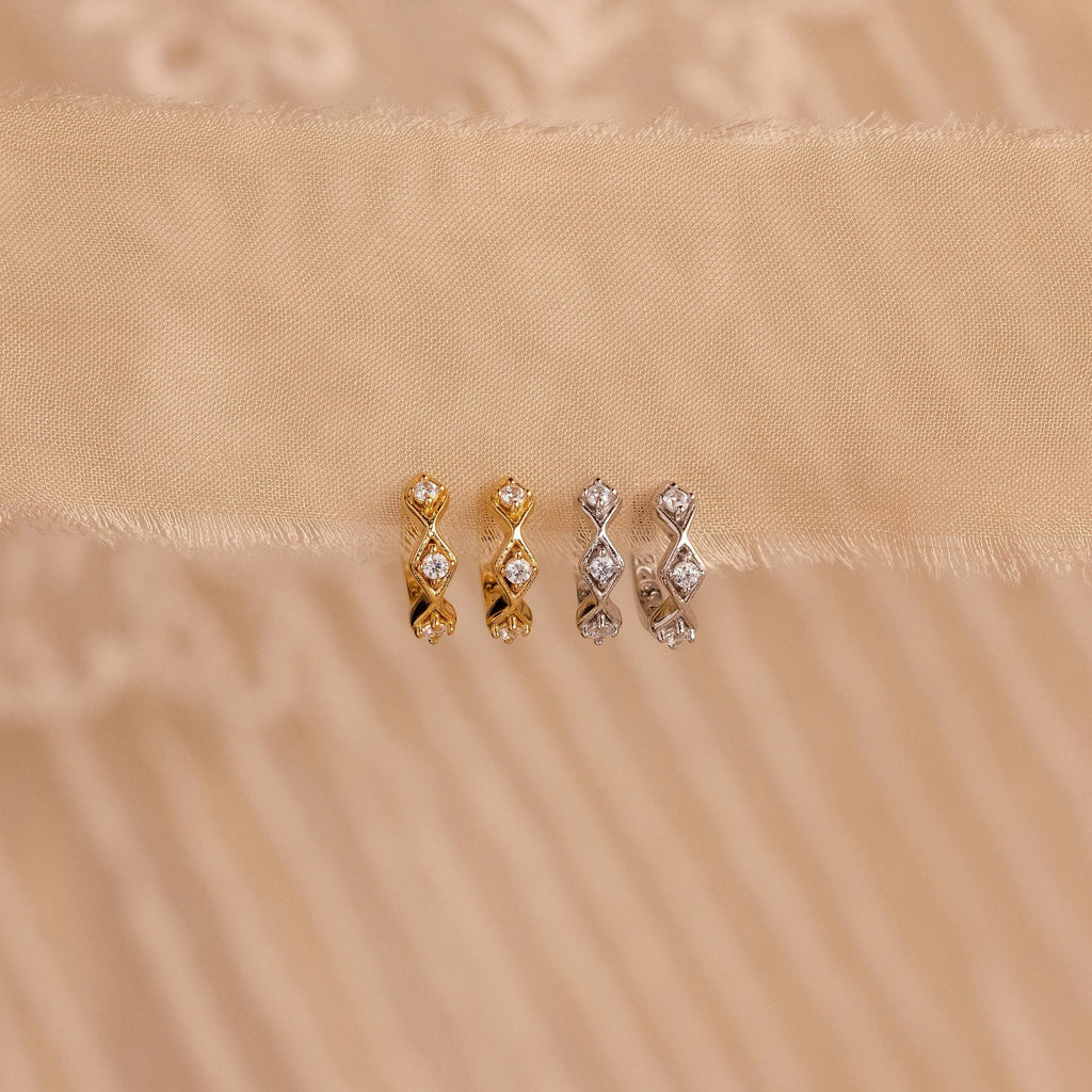 Two pairs of Ericka Diamond Tennis Huggies—one in gold, one in silver—sparkle on a beige fabric background, reflecting the timeless elegance of art deco-inspired earrings.