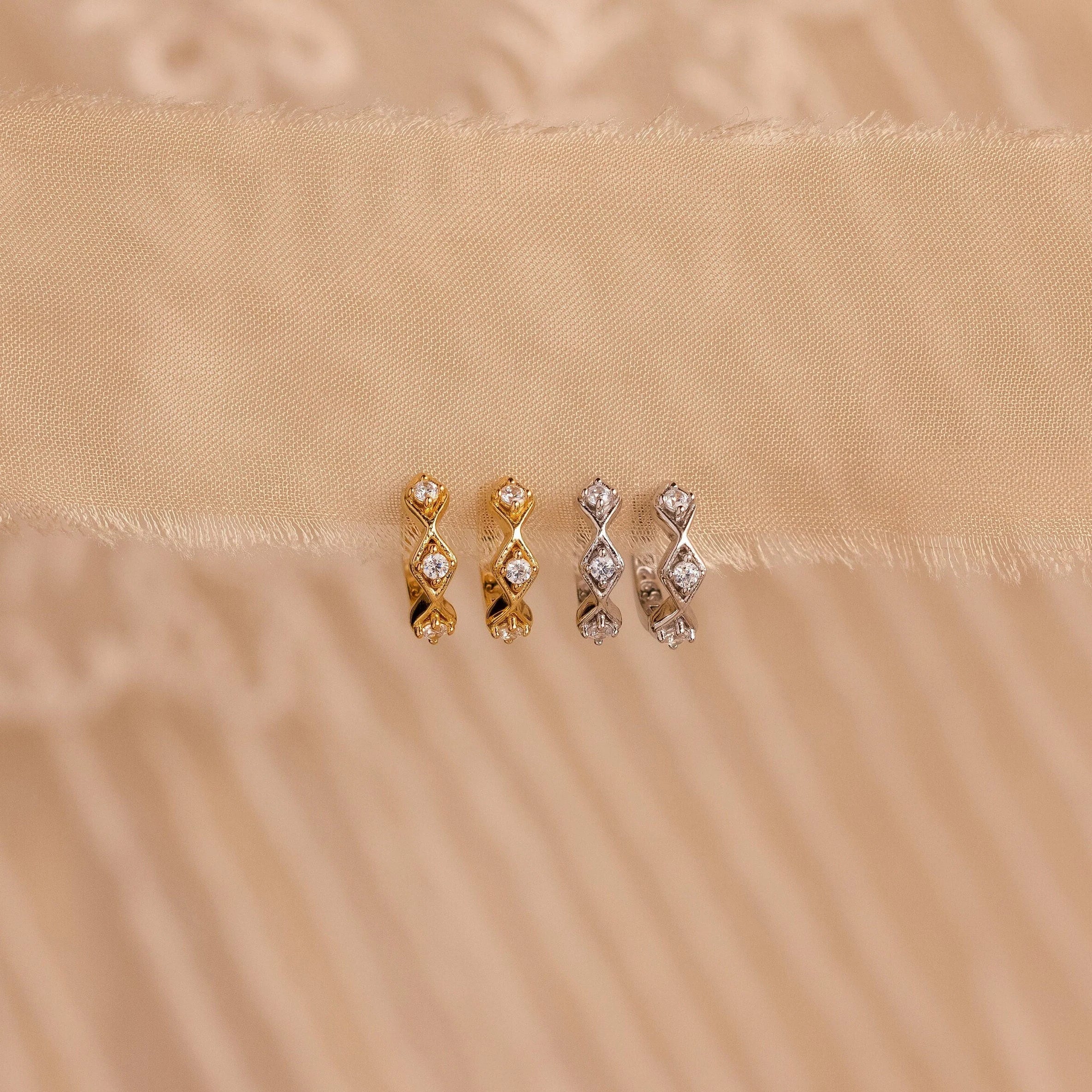 Two pairs of Ericka Diamond Tennis Huggies—one in gold, one in silver—sparkle on a beige fabric background, reflecting the timeless elegance of art deco-inspired earrings.