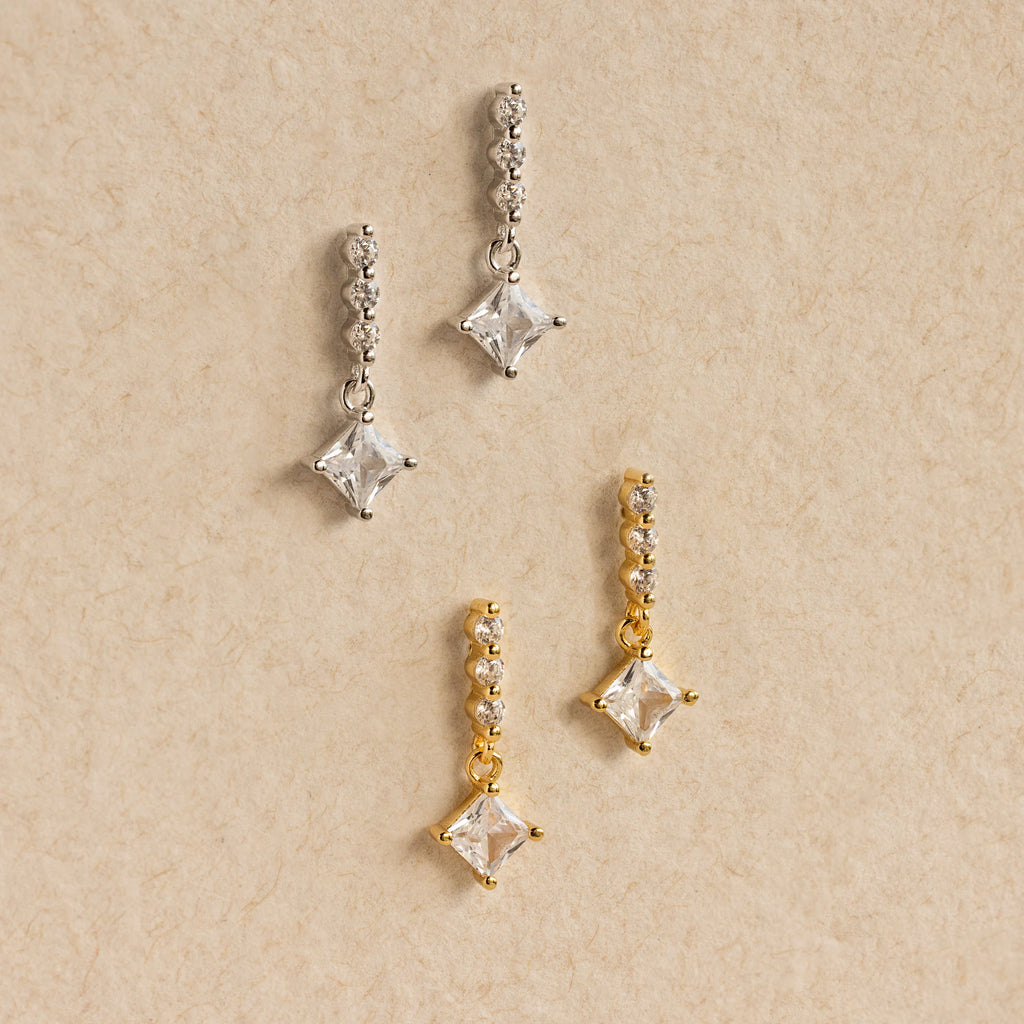 Two pairs of Princess Diamond Drop Studs—one in silver, one in gold—adorn a beige textured background, each pair featuring modern square crystals that highlight the sparkle of a princess cut gemstone.