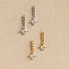 Two pairs of Princess Diamond Drop Studs—one in silver, one in gold—adorn a beige textured background, each pair featuring modern square crystals that highlight the sparkle of a princess cut gemstone.